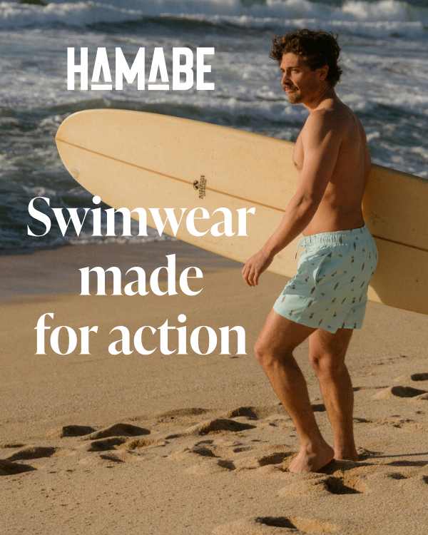 A man on the beach holding a surfboard in his hands and wearing Hamabe swim trunks.